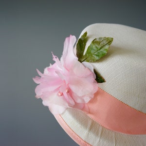 Op de afbeelding: Een witte strohoed met een roze lint en een roze bloem met groene bladeren.