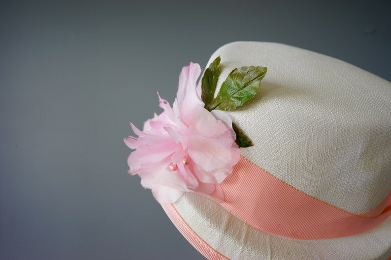 Op de afbeelding: Een witte strohoed met een roze lint en een roze bloem met groene bladeren.