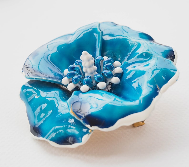 May include: A blue enamel flower brooch with a white center. The flower has a glossy finish and a slightly textured surface.