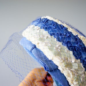May include: A vintage hat with a blue and white striped design, featuring a delicate blue netting. The hat has a light blue band and is likely made of fabric. The overall style suggests a retro or classic aesthetic.