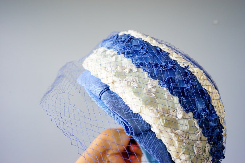 May include: A vintage hat with a blue and white striped design, featuring a delicate blue netting. The hat has a light blue band and is likely made of fabric. The overall style suggests a retro or classic aesthetic.