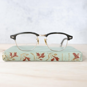 Puede incluir: Gafas con montura negra y dorada sobre un libro antiguo. Las gafas tienen un estilo clásico, con montura superior negra y bordes inferiores dorados. El libro tiene una cubierta turquesa con un diseño floral en rojo y dorado.