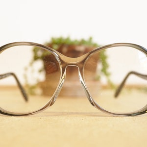 Könnte beinhalten: Eine runde, klare Brille mit einem dunklen grauen Farbton aus Kunststoff. Die Brille liegt auf einer hellbraunen Oberfläche.