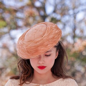 May include: A woman wearing a peach-colored feathered hat with a round crown. The hat is tilted over her forehead, and she is looking down. She is wearing a white lace dress.