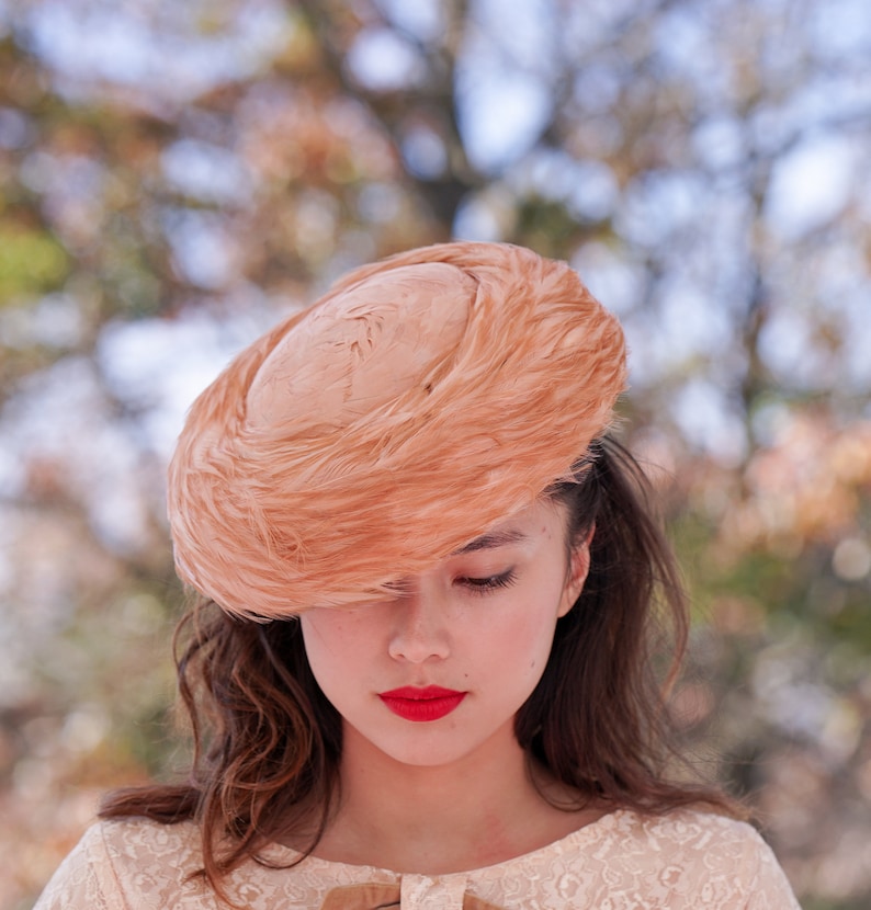 May include: A woman wearing a peach-colored feathered hat with a round crown. The hat is tilted over her forehead, and she is looking down. She is wearing a white lace dress.