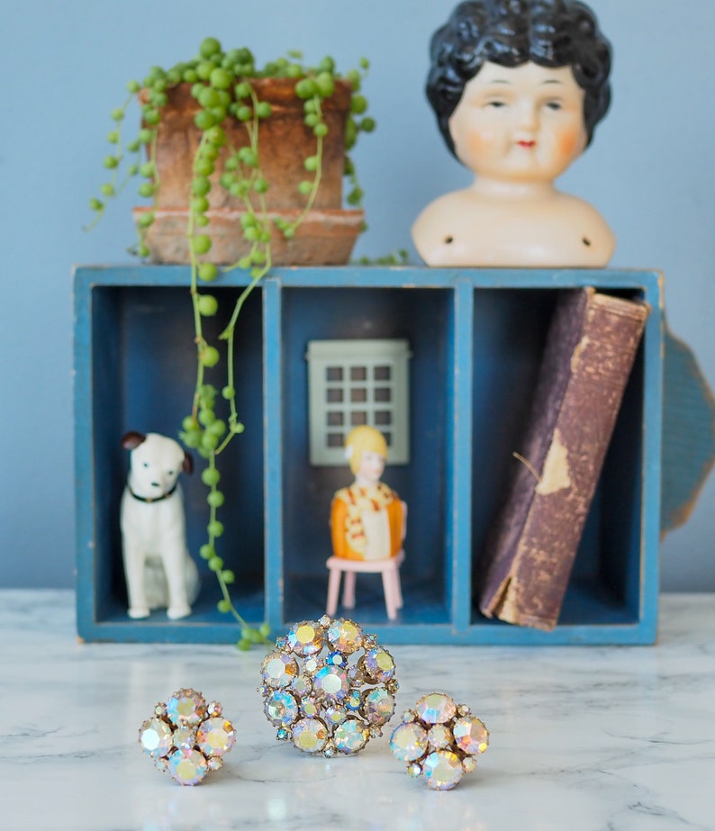 May include: A set of three iridescent rhinestone brooches in a vintage style. The brooches are round and feature a cluster of rhinestones in a variety of sizes. The brooches are displayed on a white surface in front of a blue shelf with a doll head, a potted plant, and a book.