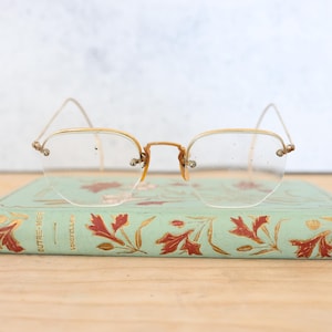 Puede incluir: Gafas vintage con montura dorada y lentes transparentes, descansando sobre un libro verde con diseños florales dorados y rojos y el texto "OUTRE-MER" y "CRISTALLYS".