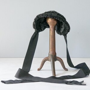 May include: A black velvet bonnet with a gathered crown and long black ribbon ties. The bonnet is displayed on a decorative stand.