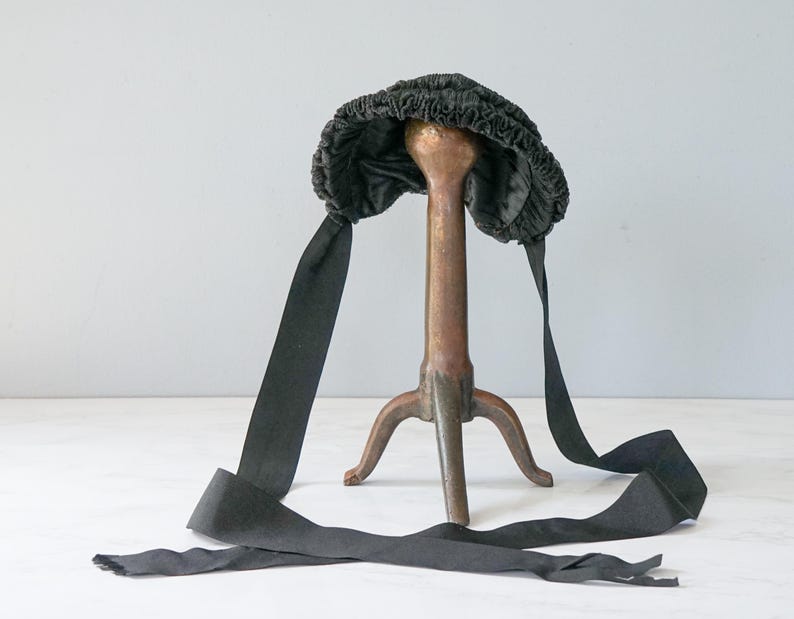 May include: A black velvet bonnet with a gathered crown and long black ribbon ties. The bonnet is displayed on a decorative stand.