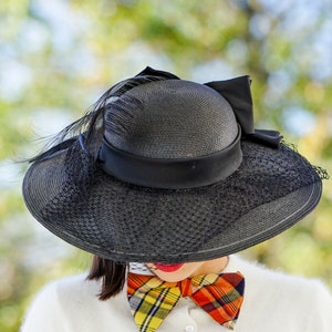 May include: A black wide-brimmed hat with a black ribbon and a feather accent. The hat is worn by a person with a plaid bow tie.