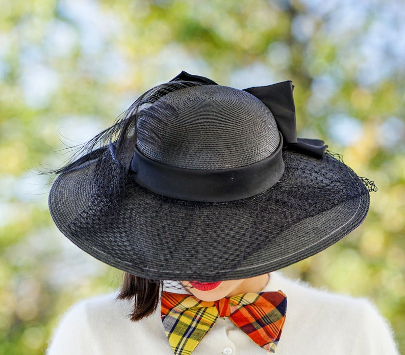 May include: A black wide-brimmed hat with a black ribbon and a feather accent. The hat is worn by a person with a plaid bow tie.
