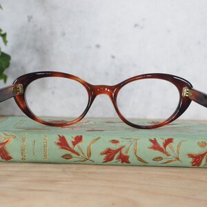 May include: A pair of tortoiseshell cat-eye glasses with gold accents. The glasses are resting on a vintage book with a green cover and gold lettering.