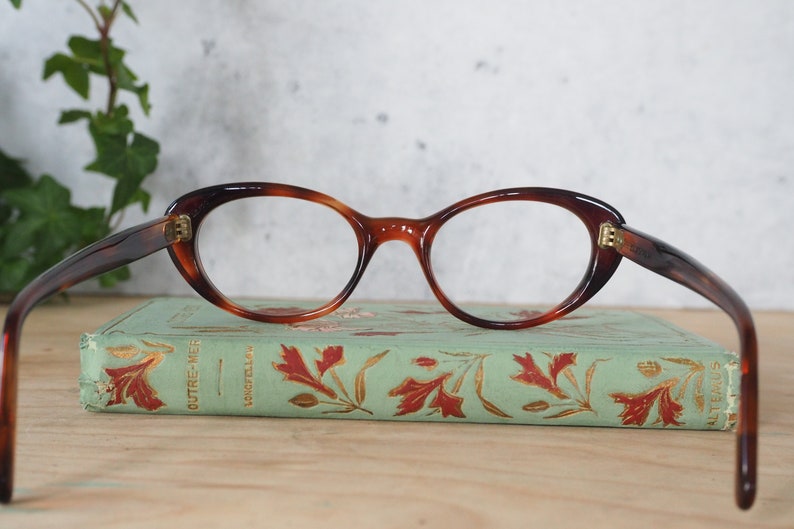 May include: A pair of tortoiseshell cat-eye glasses with gold accents. The glasses are resting on a vintage book with a green cover and gold lettering.