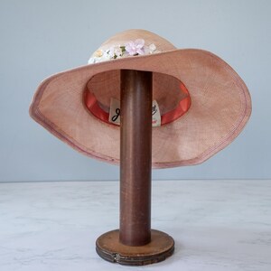 May include: A pink straw hat with a wide brim and a band of pink and white flowers. The hat is sitting on a wooden hat stand.