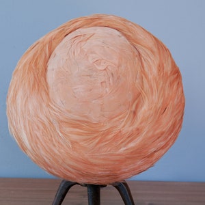 May include: A peach-colored hat made of feathers. The hat is round and has a smooth, slightly textured surface. It is sitting on a tripod stand.