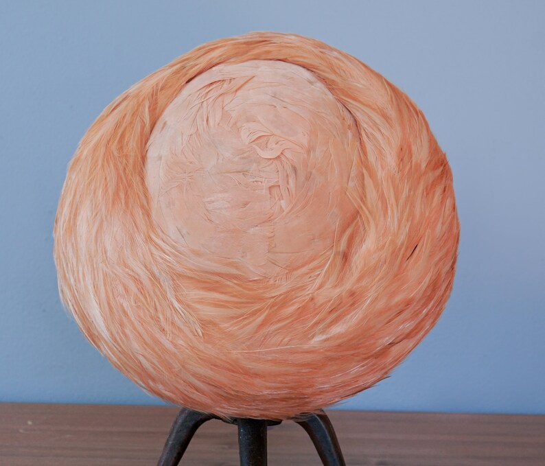 May include: A peach-colored hat made of feathers. The hat is round and has a smooth, slightly textured surface. It is sitting on a tripod stand.