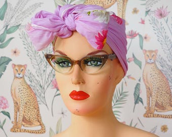 May include: A mannequin head wearing a light purple headwrap with a large knotted bow on top, featuring a floral pattern with pink and white flowers. The mannequin is also wearing brown cat-eye glasses with a gradient tint and bright red lipstick.
