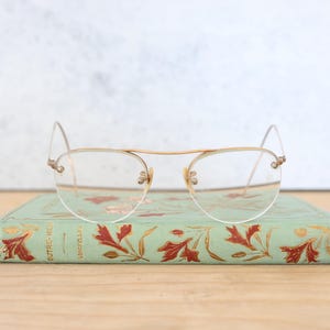 Puede incluir: Gafas transparentes con borde dorado y un diseño delicado, descansando sobre un libro vintage con una cubierta verde y ilustraciones florales rojas. El título del libro es "Unctelv" y "Autre-Mer".