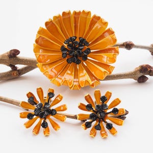 May include: A set of orange and black enamel flower-shaped jewelry. The brooch is larger than the earrings. The brooch has a center of black enamel with a flower design. The earrings have a similar design.