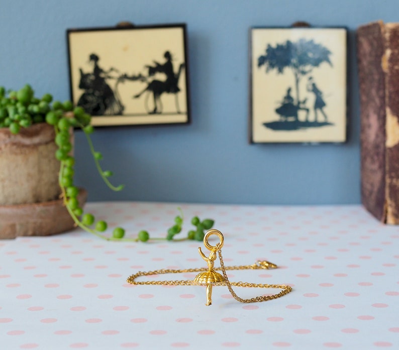 May include: A gold-colored necklace featuring a ballerina pendant. The ballerina is posed mid-leap, with arms raised. The necklace rests on a pink and white polka-dotted surface, with two framed silhouette art pieces in the background.
