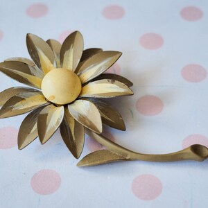 K&ouml;nnte beinhalten: Eine goldfarbene Brosche aus Metall in Form einer Blume mit gelbem Zentrum und wei&szlig;en Akzenten. Die Blume hat einen Stiel und ist wie eine G&auml;nsebl&uuml;mchen gestaltet.