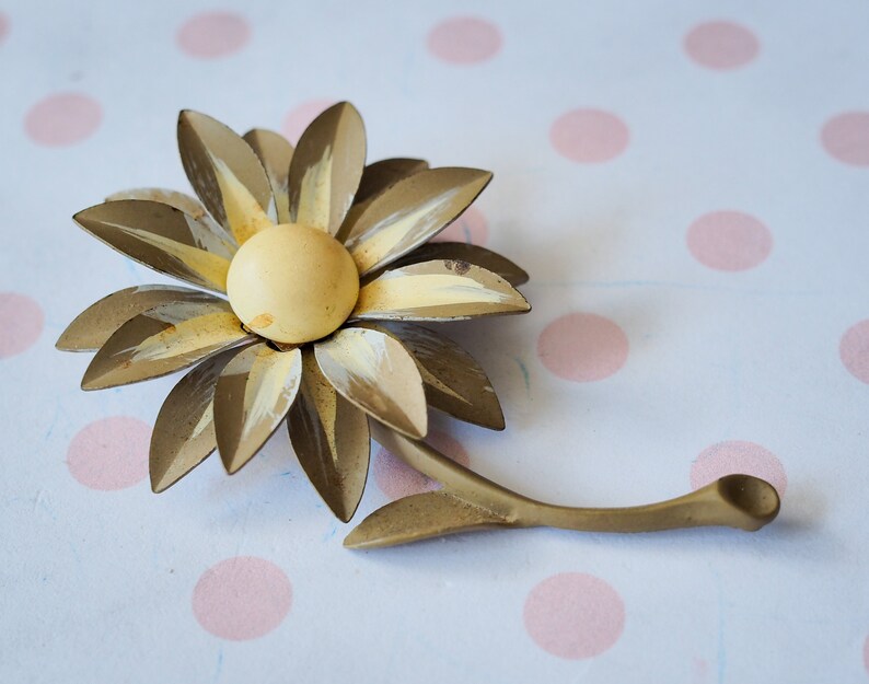 K&ouml;nnte beinhalten: Eine goldfarbene Brosche aus Metall in Form einer Blume mit gelbem Zentrum und wei&szlig;en Akzenten. Die Blume hat einen Stiel und ist wie eine G&auml;nsebl&uuml;mchen gestaltet.