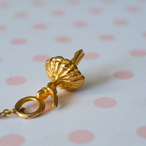 May include: A gold-colored pendant necklace featuring a ballerina in a tutu. The pendant is detailed with a textured tutu and a delicate chain. The background is a white surface with pink polka dots.
