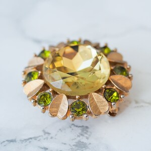 Peut inclure: Une broche dor&eacute;e avec une grande pierre de verre jaune facett&eacute; entour&eacute;e de plus petites pierres de verre vertes.