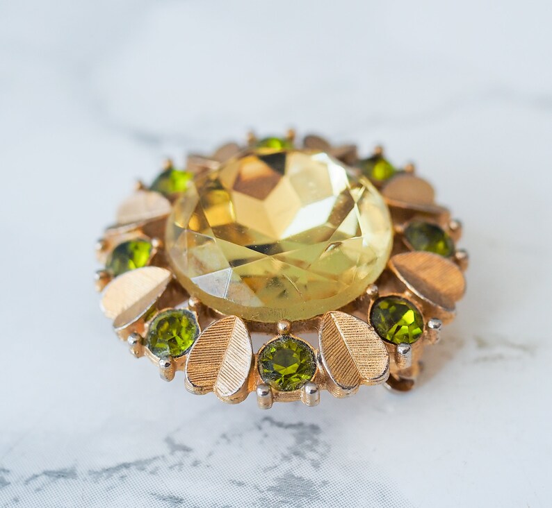 Peut inclure: Une broche dor&eacute;e avec une grande pierre de verre jaune facett&eacute; entour&eacute;e de plus petites pierres de verre vertes.