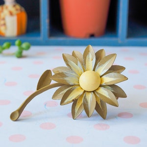 K&ouml;nnte beinhalten: Eine goldfarbene Metall-Sonnenblumenbrosche mit gelbem Zentrum. Die Sonnenblume hat eine strukturierte Oberfl&auml;che und ist mit wei&szlig;er Farbe akzentuiert.