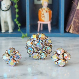 May include: Three iridescent rhinestone brooches in a cluster, flower, and circular design. The brooches are on a white surface.