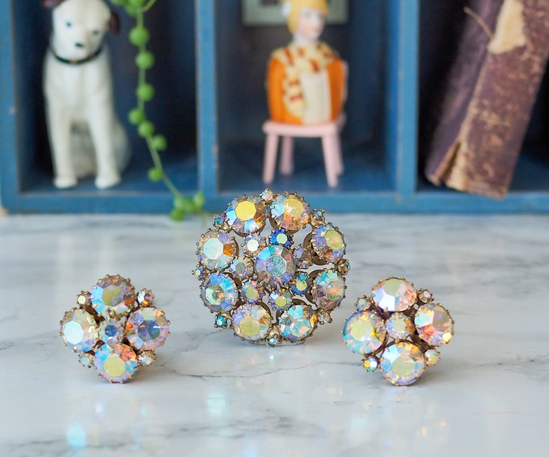May include: Three iridescent rhinestone brooches in a cluster, flower, and circular design. The brooches are on a white surface.