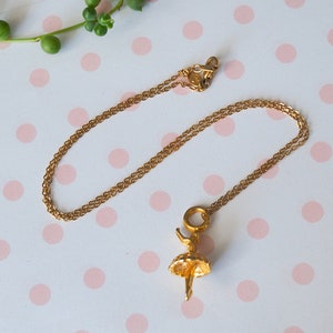May include: A gold-colored necklace with a ballerina pendant. The pendant features a detailed ballerina figure with a tutu. The chain is delicate and the clasp is visible. The necklace is displayed on a white surface with pink polka dots.