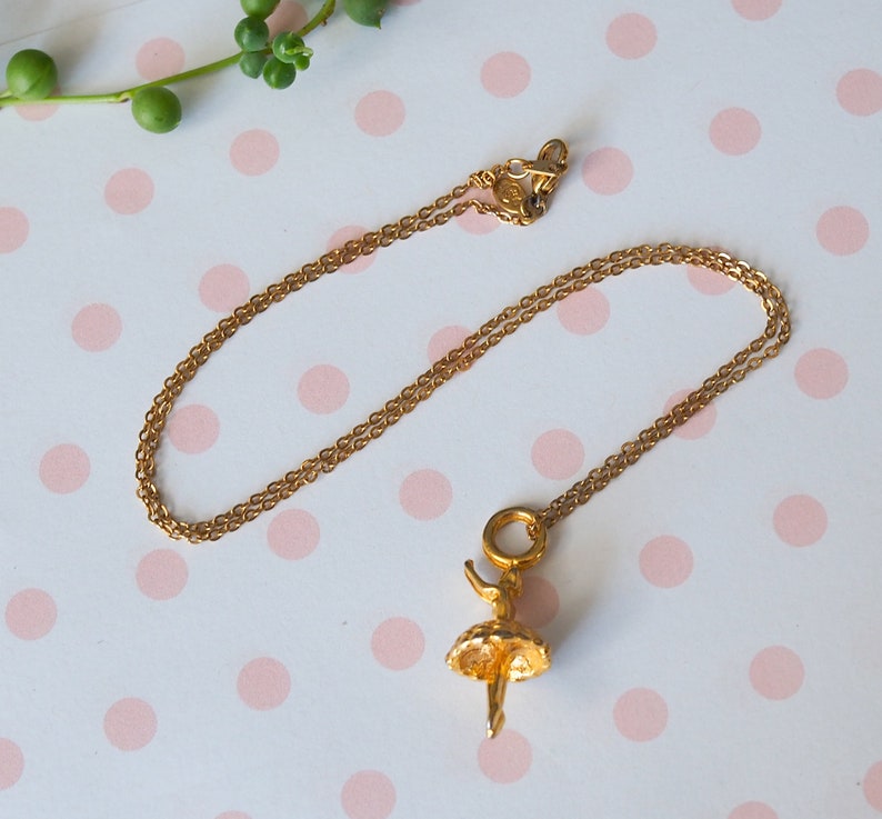 May include: A gold-colored necklace with a ballerina pendant. The pendant features a detailed ballerina figure with a tutu. The chain is delicate and the clasp is visible. The necklace is displayed on a white surface with pink polka dots.