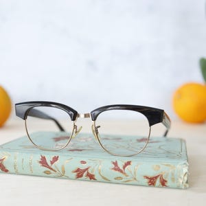 Puede incluir: Gafas con montura negra y dorada sobre un libro vintage con cubierta turquesa y diseño floral. Dos naranjas en el fondo, con una planta verde a la derecha. Las gafas tienen un estilo clásico y retro.