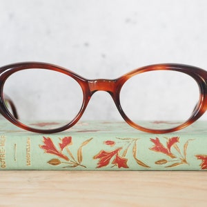 May include: A pair of tortoiseshell cat-eye glasses with clear lenses. The glasses are resting on a vintage book with a green cover and gold lettering.