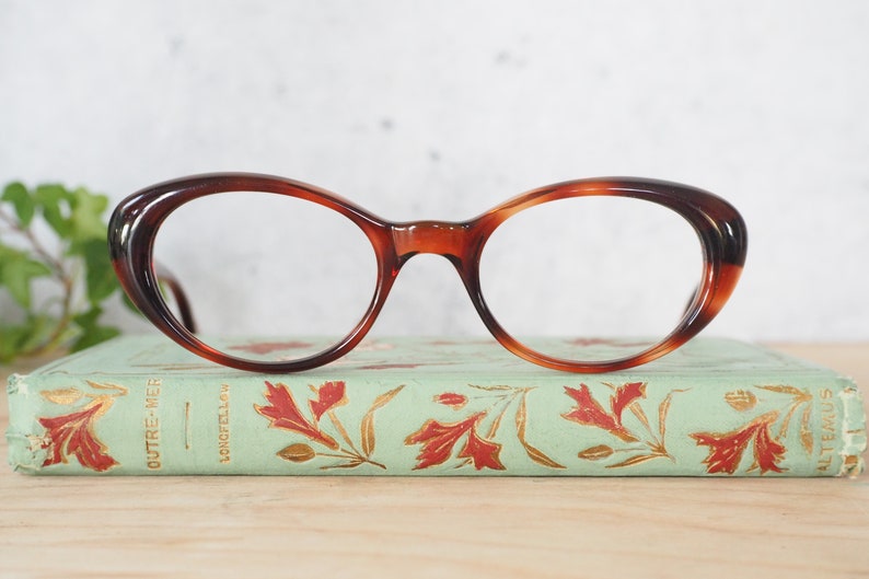 May include: A pair of tortoiseshell cat-eye glasses with clear lenses. The glasses are resting on a vintage book with a green cover and gold lettering.