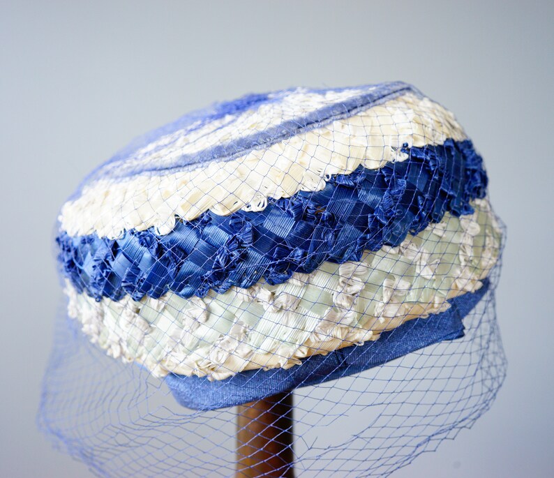May include: A vintage hat in blue, cream, and white. It has woven ribbon and netting, with a blue veil. A classic accessory.