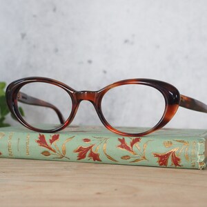 May include: Tortoiseshell cat-eye glasses with clear lenses resting on a vintage book with a green cover and gold lettering.