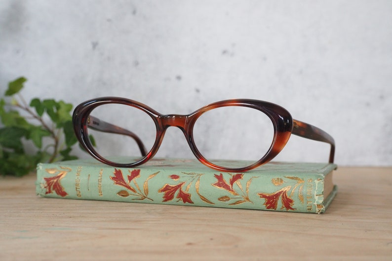 May include: Tortoiseshell cat-eye glasses with clear lenses resting on a vintage book with a green cover and gold lettering.