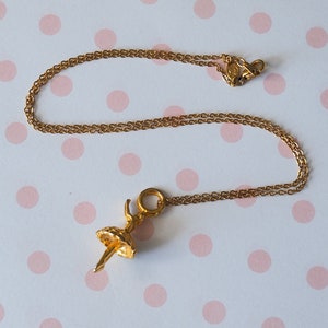 May include: A gold-colored necklace with a ballerina pendant. The pendant is a detailed figure of a ballerina in a tutu. The chain and pendant are made of a shiny, gold-toned metal. The background is a white surface with pink polka dots.