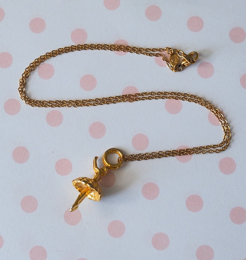 May include: A gold-colored necklace with a ballerina pendant. The pendant is a detailed figure of a ballerina in a tutu. The chain and pendant are made of a shiny, gold-toned metal. The background is a white surface with pink polka dots.