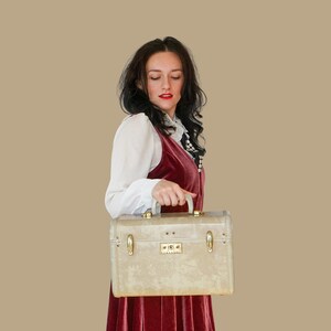 May include: A vintage beige suitcase with gold-toned hardware is held by a person. The suitcase has a rectangular shape with rounded corners and a handle on top. The person is wearing a burgundy velvet dress and a white blouse.
