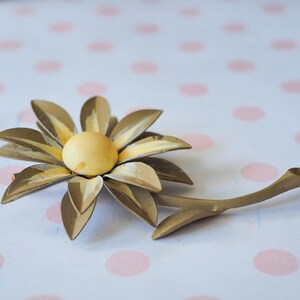 K&ouml;nnte beinhalten: Eine goldfarbene Brosche aus Metall in Form einer Blume mit gelbem Zentrum und Stiel. Die Blume hat einen Vintage-Stil.