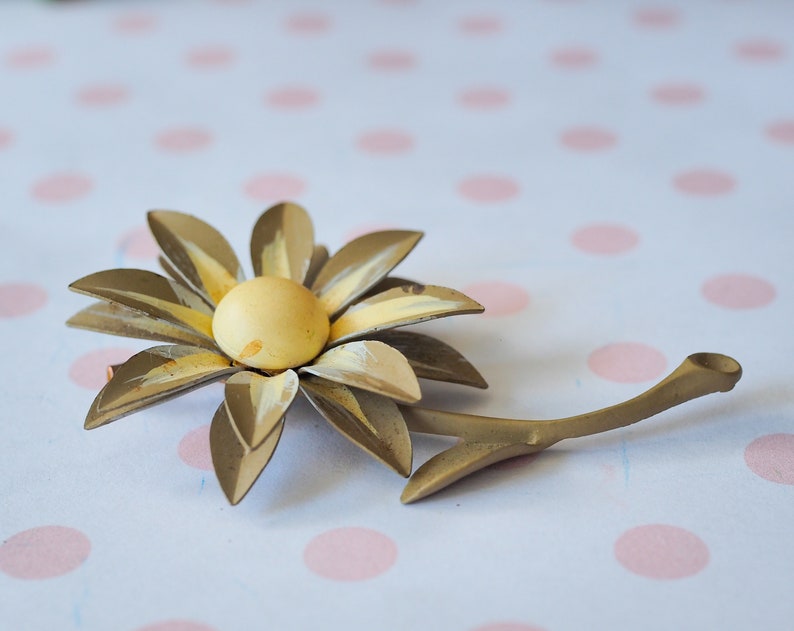 K&ouml;nnte beinhalten: Eine goldfarbene Brosche aus Metall in Form einer Blume mit gelbem Zentrum und Stiel. Die Blume hat einen Vintage-Stil.