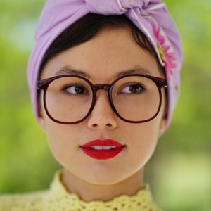 May include: A woman with dark hair wearing a purple headband with a floral design and round, dark-rimmed glasses. She is wearing a yellow crocheted top.