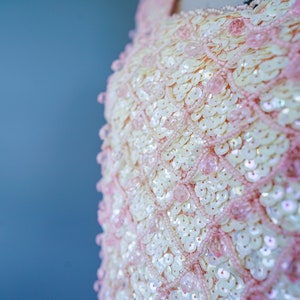 Pu&ograve; includere: Primo piano di un tessuto di paillettes rosa e bianco. Le paillettes sono disposte a formare un motivo e sono iridescenti.