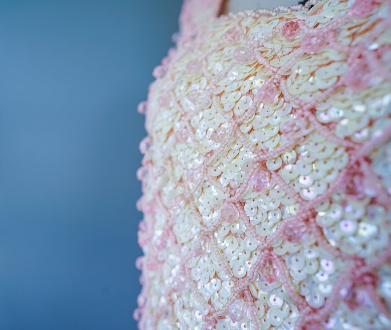 Pu&ograve; includere: Primo piano di un tessuto di paillettes rosa e bianco. Le paillettes sono disposte a formare un motivo e sono iridescenti.