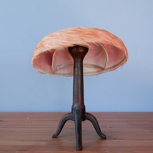 May include: A vintage hat form with a pink feather hat on top. The hat form is made of metal and has three legs.
