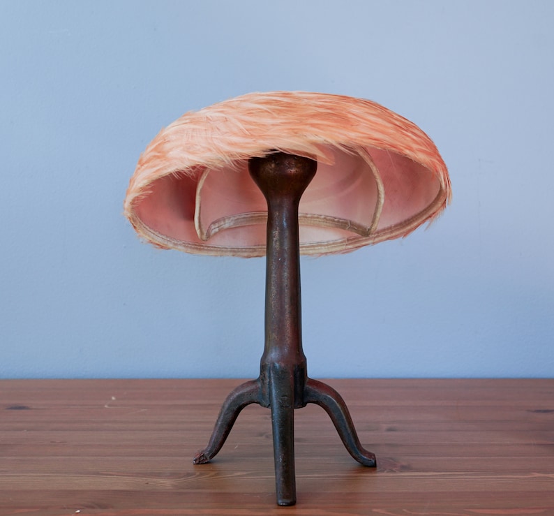 May include: A vintage hat form with a pink feather hat on top. The hat form is made of metal and has three legs.